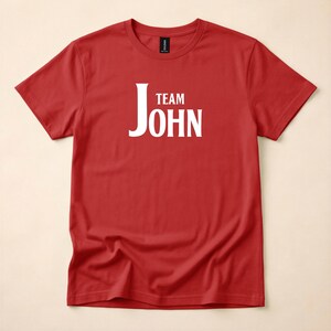 A red crewneck t-shirt featuring Team John in white text centered on the chest, laid flat against a neutral surface.