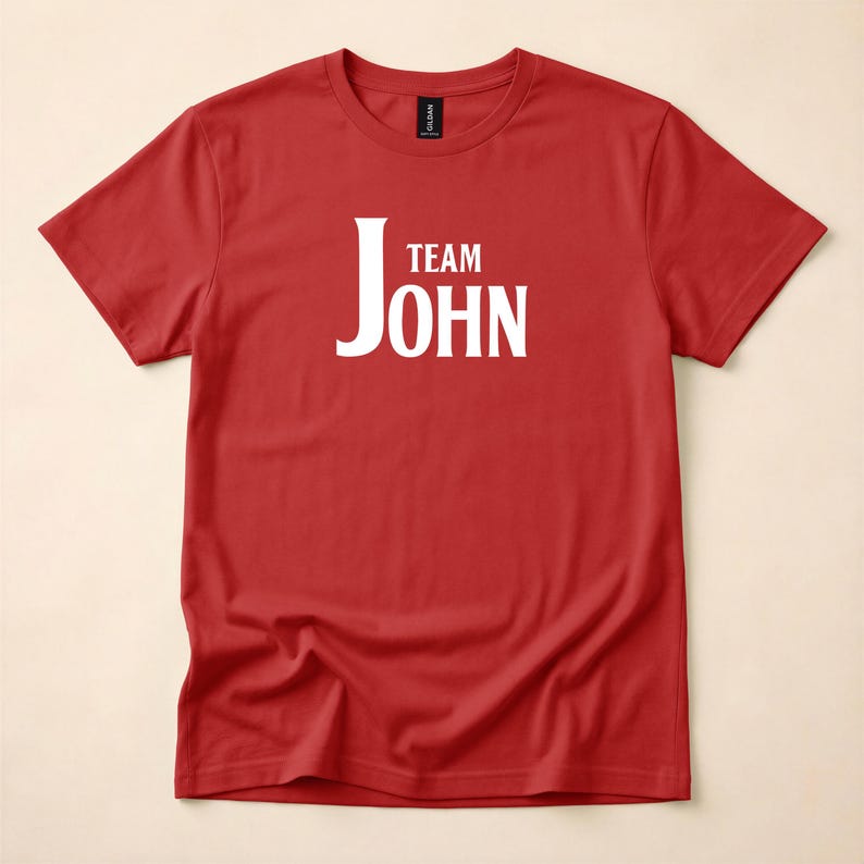 A red crewneck t-shirt featuring Team John in white text centered on the chest, laid flat against a neutral surface.