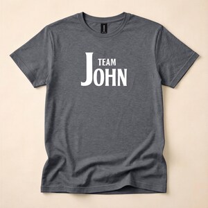 A dark gray heather t-shirt laid flat, featuring white text on the chest that reads Team John.