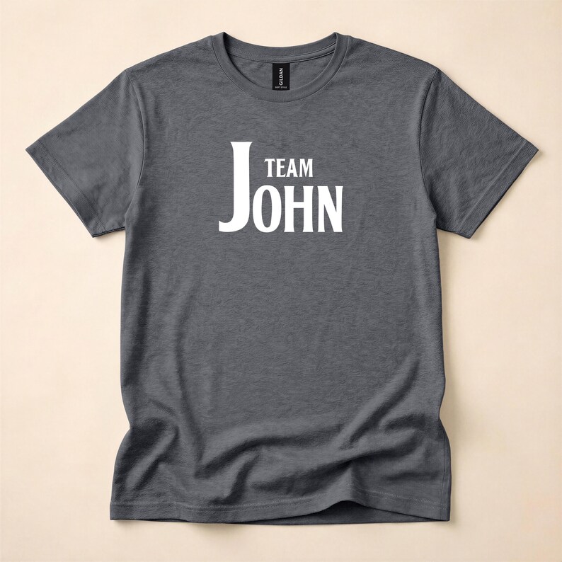 A dark gray heather t-shirt laid flat, featuring white text on the chest that reads Team John.