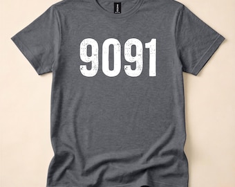 May include: A dark heather gray short-sleeve t-shirt featuring the number 9091 printed in large, white, distressed-style font across the chest. The garment has a crew neckline and a standard fit, with a visible brand label at the inner neck.