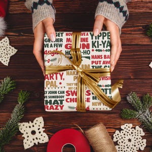 May include: A wrapped gift with a gold ribbon is held by a person. The gift wrap is cream-colored with red and green text. The gift is surrounded by Christmas decorations, including pine branches, snowflakes, and a red ribbon.
