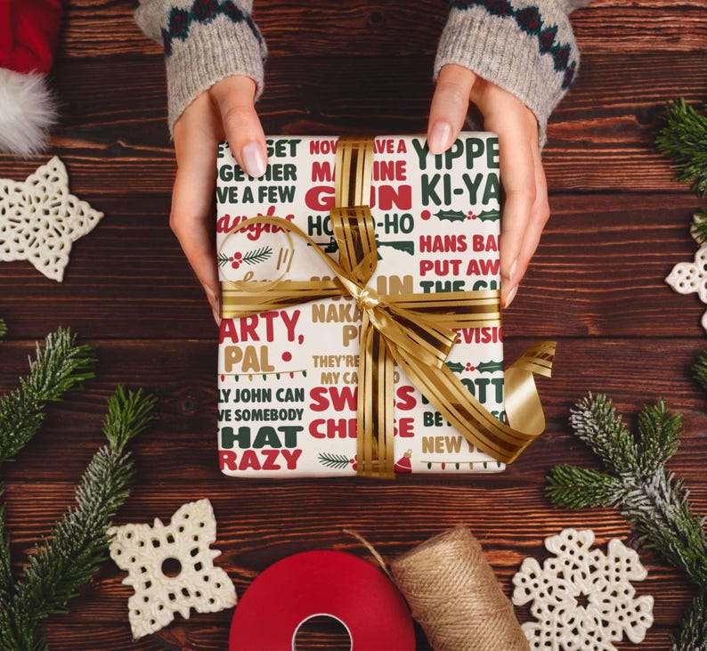 May include: A wrapped gift with a gold ribbon is held by a person. The gift wrap is cream-colored with red and green text. The gift is surrounded by Christmas decorations, including pine branches, snowflakes, and a red ribbon.