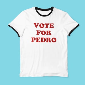 Op de afbeelding: Wit T-shirt met zwarte rand rond de kraag en mouwen. De woorden "VOTE FOR PEDRO" zijn in vette, rode letters op de voorkant gedrukt. Het shirt wordt gepresenteerd tegen een lichtblauwe achtergrond.