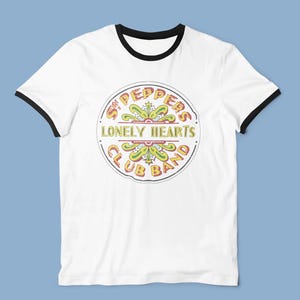 May include: White ringer t-shirt with black trim. The front displays a circular graphic with the text "Sgt. Peppers Lonely Hearts Club Band" in yellow and red, with green and yellow floral details. A classic t-shirt design.