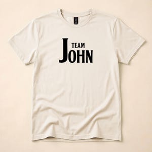 A natural-colored crewneck t-shirt with Team John printed in black text on the front, displayed flat on a light surface.