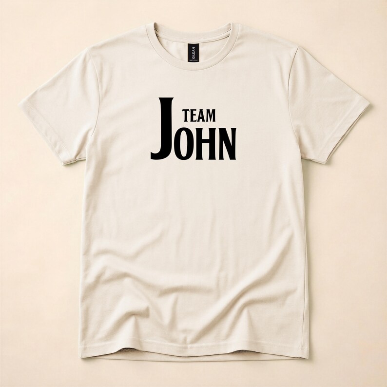 A natural-colored crewneck t-shirt with Team John printed in black text on the front, displayed flat on a light surface.