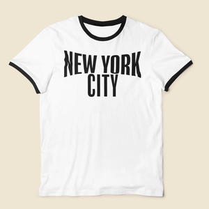 May include: White t-shirt with black trim on the collar and sleeves. The words "NEW YORK CITY" are printed in bold, black letters across the front. The shirt has a classic, casual style.