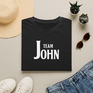 A neatly folded black t-shirt with white text reading Team John on the chest, surrounded by a straw hat, sunglasses, jeans, shoes, and small potted plants on a tan background.