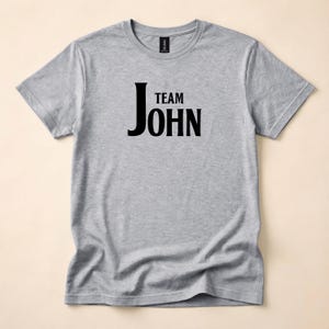 A heather grey crewneck t-shirt with Team John in black text printed on the front, laid flat against a neutral backdrop.