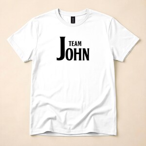 A white crewneck t-shirt with the text Team John printed in black on the chest, laid flat on a neutral background.