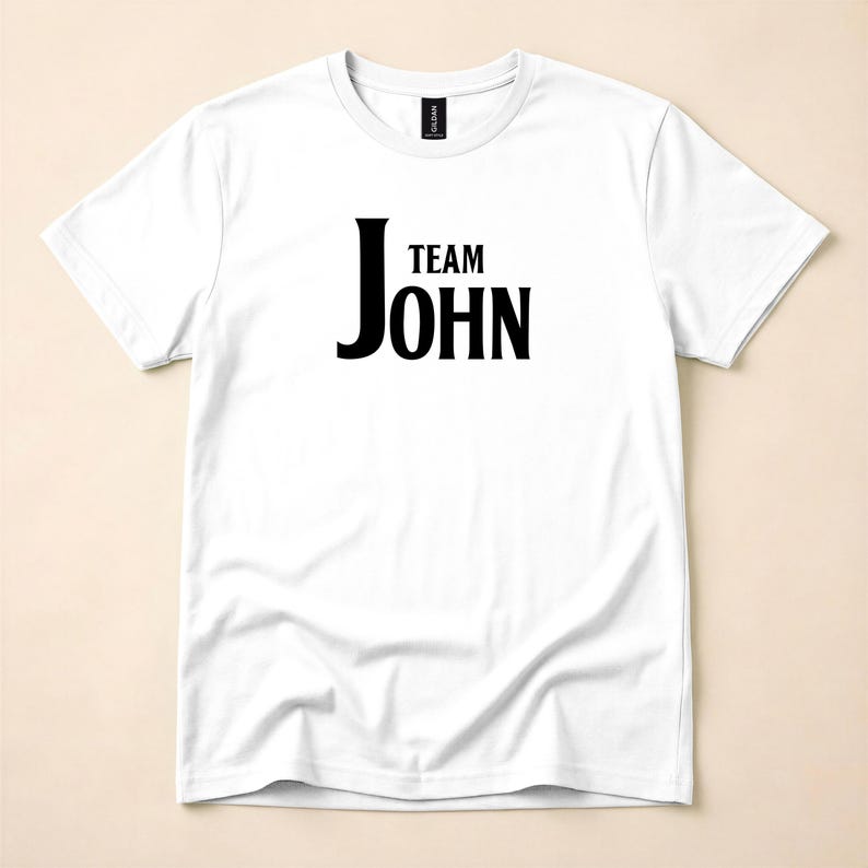A white crewneck t-shirt with the text Team John printed in black on the chest, laid flat on a neutral background.