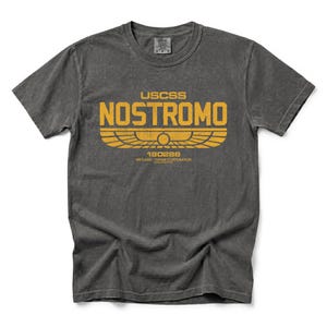 Könnte beinhalten: Ein anthrazitfarbenes Kurzarm-T-Shirt mit einer verwaschenen gelben Grafik auf der Brust. Der Aufdruck zeigt den Text USCSS NOSTROMO über einem geflügelten Emblem, darunter die Zahlen 180286 und WEYLAND-YUTANE CORPORATION.