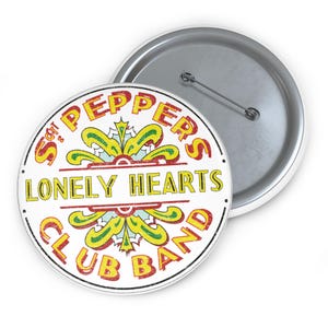 May include: A circular badge featuring the text "Sgt. Pepper's Lonely Hearts Club Band" in yellow and red on a white backdrop. It has a silver pin back and a white edge.