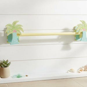Puede incluir: Un toallero decorativo con una barra de madera amarilla clara sostenida por dos recortes de palmeras. Las palmeras están pintadas en tonos de verde, azul y rosa. El toallero está montado en una pared blanca.