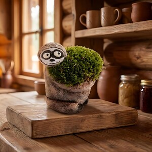 May include: A whimsical, handmade sloth planter with a mossy green plant. The planter is a light brown color with a painted sloth face. The planter sits on a wooden cutting board.