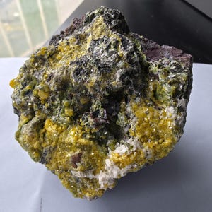 Puede incluir: Un espécimen mineral en bruto que presenta un grupo de formaciones cristalinas de color amarillo vibrante incrustadas en una matriz rocosa oscura y texturizada. La superficie muestra una mezcla de depósitos minerales amarillo brillante, verde oliva y gris carbón.
