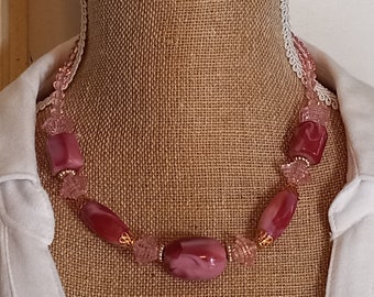 Peut inclure: Un collier de perles roses avec une variété de formes et de tailles. Le collier présente de grandes perles rectangulaires et ovales dans des tons de rose et de bordeaux, avec de plus petites perles roses claires et des accents dorés.