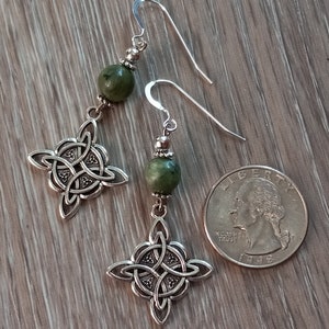 May include: A pair of silver-toned Celtic knot earrings with green stone beads. The earrings are displayed next to a U.S. quarter for size comparison. The earrings have a hook closure.