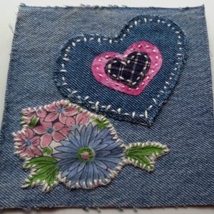 May include: A denim square with two fabric appliqués. One is a blue heart with a pink and plaid heart inside, stitched with white thread. The other is a floral design with pink, blue, and green fabric, also stitched with white thread.