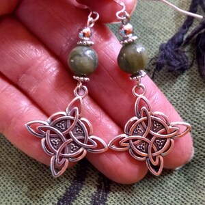 May include: Silver-toned Celtic knot earrings with olive green stone beads. The earrings feature a four-leaf clover design with intricate knotwork. The earrings have a hook closure and are displayed against a green background.
