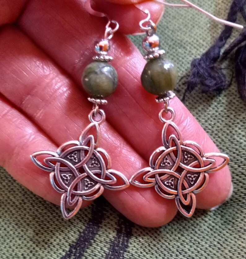 May include: Silver-toned Celtic knot earrings with olive green stone beads. The earrings feature a four-leaf clover design with intricate knotwork. The earrings have a hook closure and are displayed against a green background.