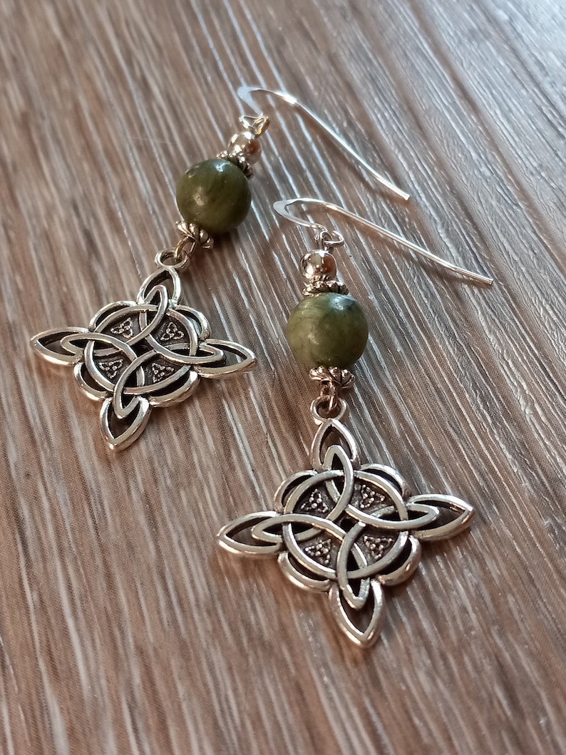 May include: A pair of silver-toned Celtic knot drop earrings. Each earring features a green bead, silver accents, and a four-pointed knot design. The earrings hang from silver ear wires.