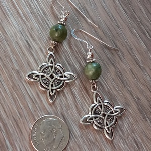 May include: A pair of silver-toned Celtic knot earrings with green stone beads. The earrings feature intricate knotwork designs and are displayed next to a U.S. dime for size comparison. The earrings hang from silver hooks.