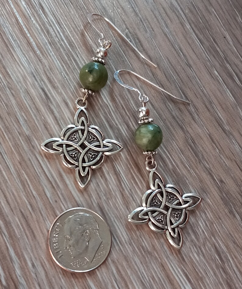 May include: A pair of silver-toned Celtic knot earrings with green stone beads. The earrings feature intricate knotwork designs and are displayed next to a U.S. dime for size comparison. The earrings hang from silver hooks.