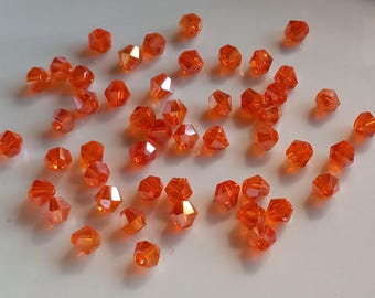 Puede incluir: Un conjunto de cuentas facetadas de color naranja dispersas sobre una superficie clara. Las cuentas tienen un aspecto brillante, similar al de una gema, con múltiples facetas que capturan la luz. Adecuado para la fabricación de joyas y manualidades.