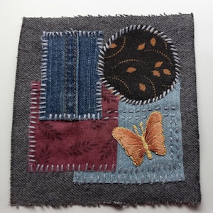 May include: A square textile art piece with a gray border. It features patches of denim, burgundy fabric with a leaf pattern, and light blue fabric. An embroidered orange butterfly is on the light blue patch. A black circle with a gold leaf pattern is also present.