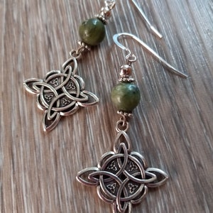 May include: A pair of silver-toned dangle earrings. Each earring features a Celtic knot design and a green, round bead. The earrings have a hook closure. The earrings are displayed on a wooden surface.