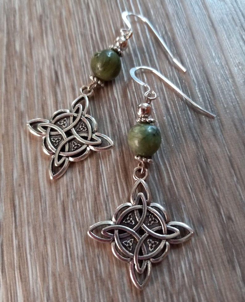 May include: A pair of silver-toned dangle earrings. Each earring features a Celtic knot design and a green, round bead. The earrings have a hook closure. The earrings are displayed on a wooden surface.
