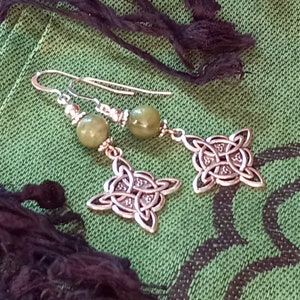 May include: Silver-toned Celtic knot earrings with olive green beads. The earrings feature a drop design with intricate knotwork pendants and a hook closure. The background is a textured green fabric.