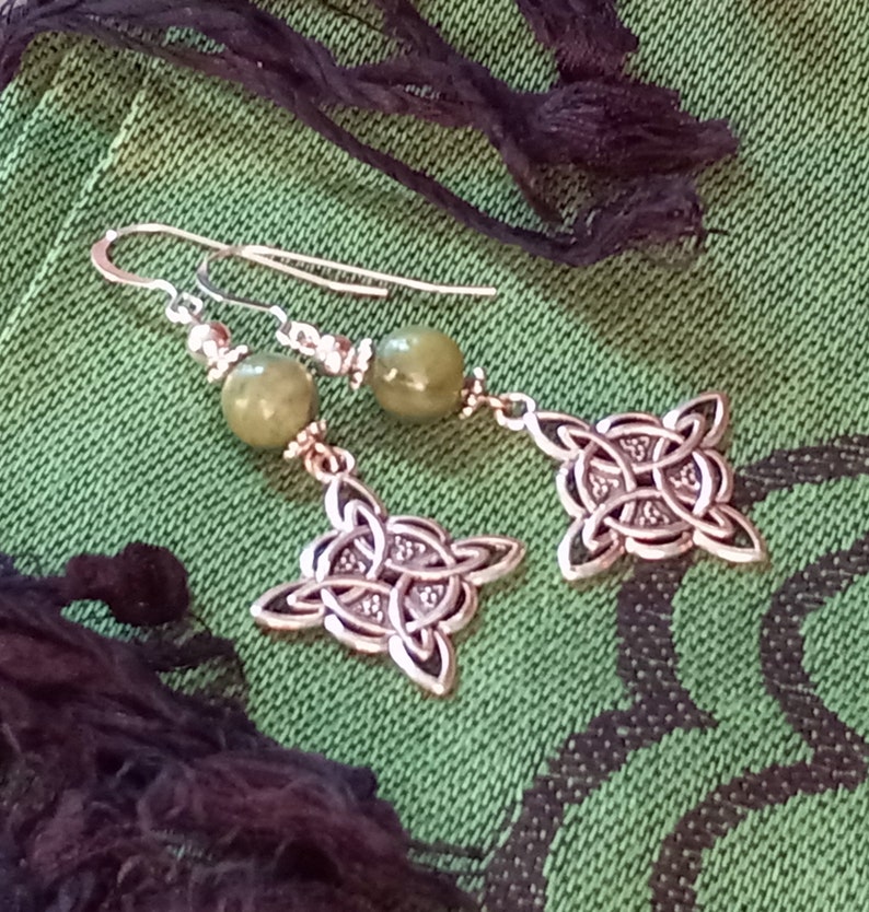 May include: Silver-toned Celtic knot earrings with olive green beads. The earrings feature a drop design with intricate knotwork pendants and a hook closure. The background is a textured green fabric.