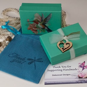 May include: An overhead shot showcases a collection of teal gift boxes, a teal drawstring bag, and a pearl necklace. The boxes feature a hummingbird and floral design. The drawstring bag has the text "Thank you!" and a dragonfly graphic. A wooden heart charm adorns one box.