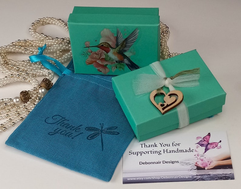 May include: An overhead shot showcases a collection of teal gift boxes, a teal drawstring bag, and a pearl necklace. The boxes feature a hummingbird and floral design. The drawstring bag has the text "Thank you!" and a dragonfly graphic. A wooden heart charm adorns one box.