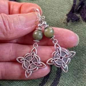 May include: A pair of silver-toned earrings featuring a Celtic knot design. Each earring has a green, round bead at the top, with a decorative silver element and a Celtic knot pendant. The earrings are held up by a clear hook.