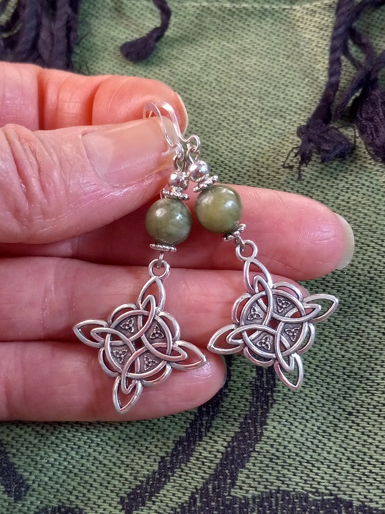 May include: A pair of silver-toned earrings featuring a Celtic knot design. Each earring has a green, round bead at the top, with a decorative silver element and a Celtic knot pendant. The earrings are held up by a clear hook.