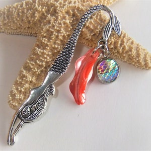 May include: A silver mermaid tail bookmark with a red coral charm and a round iridescent charm. The bookmark is displayed on a starfish.