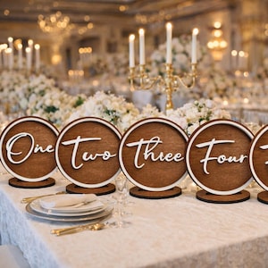 May include: Wooden table number signs with white script from one to six. The round signs have a dark brown center and a white border, set on a white tablecloth with gold accents. The background includes floral arrangements and candles.