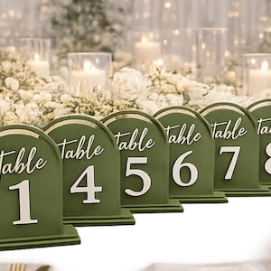 May include: Olive green table number signs with gold trim and white lettering. Each sign is arched at the top and displays the word "Table" and a number from 1 to 8. The signs are arranged in a row, suitable for a wedding or event.