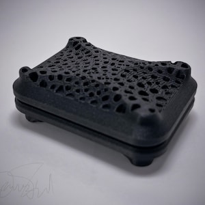 May include: A black, square box with a textured, openwork lid. The box has rounded corners and sits on four small feet. The lid features a complex, organic pattern of holes and lines. The box appears to be 3D-printed.