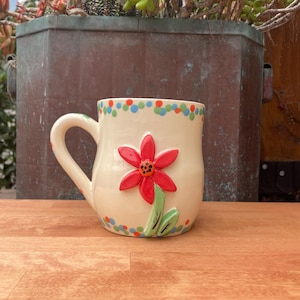 May include: Cream-colored ceramic mug with a red flower and green leaves. The mug has a handle and is decorated with colorful polka dots around the rim. The mug is sitting on a wooden surface.