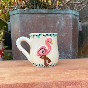 Op de afbeelding: Crèmekleurige keramische mok met een roze flamingo-ontwerp. De flamingo heeft een gele snavel en bruine poten. De rand en het handvat zijn versierd met blauwe en groene stippen. Een charmante toevoeging voor elke keuken.