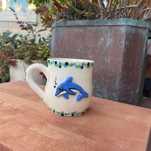 Może przedstawiać: Kremowy ceramiczny kubek z niebieskim wzorem delfina. Kubek ma zakrzywiony uchwyt i jest ozdobiony niebieskimi i zielonymi kropkami wokół krawędzi. Delfin to aplikacja 3D. Kubek stoi na drewnianej powierzchni.