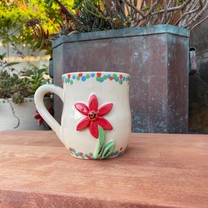 May include: A cream-colored ceramic mug with a red flower design and green leaves. The mug has a white handle and is decorated with colorful polka dots around the rim. The mug is sitting on a wooden surface.