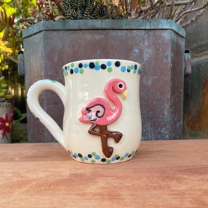 Op de afbeelding: Crèmekleurige keramische mok met een roze flamingo-ontwerp. De flamingo heeft een zwarte omtrek en een gele snavel. De rand is versierd met blauwe en groene stippen. Het handvat is wit.