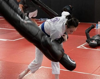Könnte beinhalten: Eine Person in einem weißen Kampfsportanzug übt eine Grappling-Technik mit einer schwarzen Trainingspuppe. Der Trainingsbereich hat rote Matten und weiße Linien. Andere Personen sind im Hintergrund.