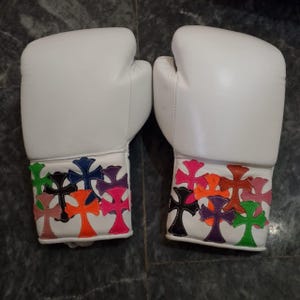 May include: A pair of white boxing gloves with colorful cross designs on the wrist cuffs. The crosses are in various colors, including pink, orange, green, and purple. The gloves are made of a smooth material.